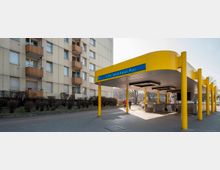 U-Bahn-Eingang des Bahnhofs Jakob-Kaiser-Platz in Berlin, mit einem gelben Dach und gelben Säulen, daneben ein Wohngebäude mit Balkonen im Hintergrund. Auf dem Vorplatz befinden sich Gehwege, Büsche und Passanten.