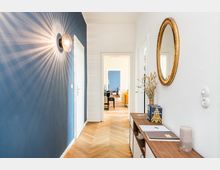 Heller Flur in einer Wohnung mit blau gestrichener Akzentwand, Wandlampe mit Strahlenlicht, weiß gestrichenen Türen und Fischgrätparkett. An der rechten Wand hängen ein runder Spiegel mit Goldrahmen und darunter steht ein schmaler Konsolentisch mit Deko; im Hintergrund führt der Flur in einen Wohnraum.