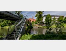 Moderne Fußgängerbrücke mit einer Treppe aus Glas und Metall über einen Fluss, umgeben von grüner Vegetation. Im Hintergrund sind eine Kirche mit rotem Dach und Glockenturm sowie ein mehrstöckiges Wohngebäude sichtbar.