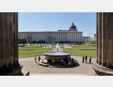 Blick durch Säulen auf ein Freigelände mit symmetrischen Rasenflächen, einem zentralen Brunnen und im Hintergrund das Humboldt Forum, ein großes historisch anmutendes Gebäude mit Kuppel. Im Vordergrund befindet sich eine große steinerne Schale, um die Menschen sitzen.