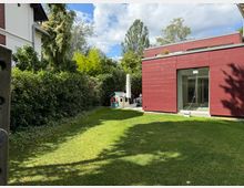 Ein moderner Garten mit einer roten, kubischen Hausfassade auf der rechten Seite, großen Glasfenstern und einer gepflegten Rasenfläche. Links befinden sich hohe Hecken und Bäume, während im hinteren Bereich ein kleines Spielhaus und eine Terrasse mit Sonnenschirm zu sehen sind.