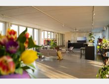 Ein moderner, lichtdurchfluteter Raum mit großen Fensterfronten, minimalistischer Möblierung und grünen Pflanzen. Im Vordergrund stehen bunte Tulpen, während im Hintergrund Sitzmöbel, ein Tresen und ein Kleiderständer mit farbenfrohen Kleidungsstücken zu sehen sind.