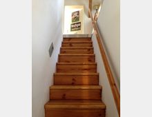 Treppenaufgang aus Holz in einem Innenraum mit weiß gestrichenen Wänden; an der rechten Seite ein Holzhandlauf, am oberen Ende der Treppe ein beleuchteter Flur mit Postern an der Wand.