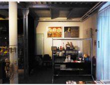 Ein Innenraum mit industrieller Ästhetik, erkennbar durch sichtbare Stahlträger und eine Betondecke. Der Raum enthält eine Vitrine mit Büchern und Gegenständen, im Hintergrund hängen Plakate und Bilder an einer weißen Wand. Links sind an einem Pfosten Puppen aufgehängt.