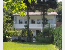 Zweistöckiges weißes Haus mit Flachdach und umlaufendem Balkon im oberen Stockwerk, umgeben von Bäumen und dichtem Grün. Im Vordergrund ist ein Garten mit Rasenfläche, Büschen und kleinen Laternen sichtbar.