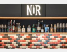 Innenansicht einer Bar mit einer langen Holzablage, auf der zahlreiche Spirituosenflaschen vor einer dunklen Wand stehen. Unter der Ablage befindet sich eine Wandverkleidung aus rechteckigen, mehrfarbigen Kachelsteinen in Rot-, Grau- und Beigetönen.