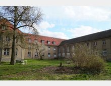 Ein rechteckiger Innenhof mit grünem Rasen und vereinzelten Bäumen, umgeben von einem mehrstöckigen Gebäude mit beigen Wänden und roten Ziegeldächern. Im Vordergrund stehen einfache Gartenmöbel, und im Hintergrund sind Fenster mit weißen Rahmen zu sehen.