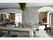 Gemütliches Café mit rustikaler Einrichtung, einschließlich Steinwänden, gewölbten Türdurchgängen und einem barähnlichen Tresen mit Mosaikfliesen. Die Sitzbereiche sind mit Holztischen und -stühlen ausgestattet, und im Hintergrund sind Fenster mit Blick nach draußen zu sehen.