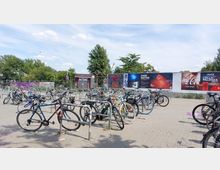 Ein Fahrradabstellplatz mit mehreren geparkten Fahrrädern auf einem gepflasterten Platz. Im Hintergrund sind Bäume, eine rot-weiße Fotoautomat-Kabine und eine Plakatwand mit verschiedenen Werbeanzeigen zu sehen.