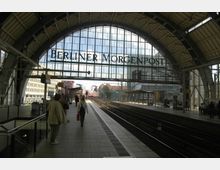 Ein moderner Bahnhof mit einem großen Glasdach und einer Stahlkonstruktion, auf dem die Schrift "Berliner Morgenpost" prangt. Im Vordergrund befinden sich Bahnsteige mit wartenden Personen, während im Hintergrund Gleise in die Ferne führen.