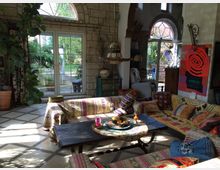 Wohnzimmer mit ethnisch inspiriertem Dekor, niedrigen Sofas mit bunten, gemusterten Decken und Kissen. Große Fenster mit Blick auf einen Garten, Steinwände und Pflanzen im Innenraum schaffen eine helle und einladende Atmosphäre.