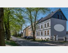 Straßenansicht mit gepflastertem Weg und Bäumen, die Schatten spenden. Rechts steht ein zweistöckiges Gebäude mit grauer Fassade, weißen Verzierungen um die Fenster und einem kleinen Hof, der durch ein schmiedeeisernes Tor begrenzt ist. Links parken Autos entlang der Straße.