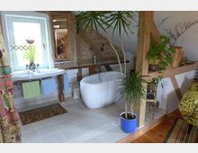 Dachgeschoss-Badezimmer mit freistehender, weißer Badewanne, Waschtisch mit Spiegel und einem kleinen Fenster. Sichtbare Holzbalken, Backsteinwand und mehrere Pflanzen in Töpfen schaffen eine rustikale Atmosphäre.