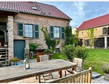 Backsteinbauernhaus mit roten Ziegeldächern, grünen Fensterläden und rankendem Efeu an der Fassade. Im Vordergrund ein möblierter Hof mit Holztisch und Stühlen, daneben gepflegter Garten mit Rasen und Blumentöpfen.