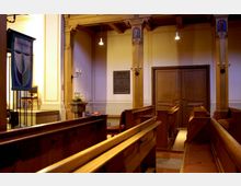 Innenraum einer kleinen Kirche oder Kapelle mit Holzbänken, verzierten Säulen, schlichten Wandleuchtern und einem Holztürportal. Im Vordergrund sind eine Vase mit Blumen und ein dekoratives Kirchenbanner zu sehen.