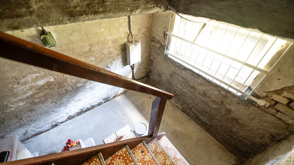 Blick in einen alten Keller- oder Treppenhausbereich mit roh verputzten, teils freiliegenden Ziegelwänden und einer Holztreppe mit gemusterten Fliesenstufen. Rechts fällt Tageslicht durch ein vergittertes Fenster, an der Wand hängen ein kleiner Elektroverteiler und Leitungen.