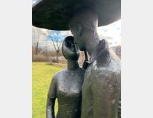 Nahaufnahme einer Bronzeskulptur im Freien, die zwei stehende Personen darstellt, die einander zugewandt sind, teilweise von einem Dach oder Schirm bedeckt. Im Hintergrund ist eine grasbewachsene Fläche, kahle Bäume und ein bewölkter Himmel zu sehen.