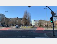 Straßenkreuzung in einer städtischen Umgebung mit Zebrastreifen und Radwegen in rot markiert. Im Hintergrund sind mehrstöckige Wohn- und Bürogebäude zu sehen, umgeben von kahlen Bäumen und einer grünen Verkehrsinsel.