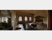 Ein historisch anmutender Saal mit hohen Decken, mehreren Fenstern mit roten Vorhängen und einer Bühne im Hintergrund. Im Raum befinden sich Stühle und Tische, teils in unaufgeräumter Anordnung, sowie ein Kronleuchter an der Decke.