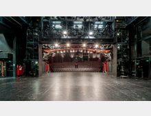 Blick von der Bühne eines modernen Theatersaals in den Zuschauerraum mit gestaffelten Sitzreihen und rotem Geländer. Oberhalb befinden sich zahlreiche Scheinwerfer und technische Konstruktionen; die Seiten sind von schwarzer Bühnentechnik eingerahmt.