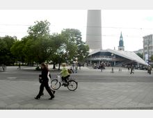 Straßenszene auf einem Platz in Berlin mit Bäumen, Passanten und Radfahrenden im Vordergrund. Im Hintergrund sind der Fernsehturm, ein modernes Gebäude mit breiten Treppen und die Spitze einer Kirche sichtbar.