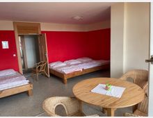 Ein einfaches Gästezimmer mit roten Wänden, drei Betten mit Holzrahmen, einem runden Holztisch und vier Korbstühlen. Im Hintergrund ist eine offenstehende Tür zu einem angrenzenden Raum sichtbar.