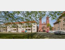 Mehrfamilienhäuser mit hellen Fassaden und roten Ziegeldächern stehen neben einer roten Backsteinkirche mit zwei Türmen und spitzem Giebel. Im Vordergrund sind ein umzäunter Rasenbereich, parkende Autos und Bäume mit frischem Laub zu sehen.