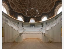 Runder historischer Saal mit einer Kuppeldecke und filigranen Holzgeländern, umgeben von oval angeordneten Balkonen. Hohe Fenster lassen Tageslicht herein, während die Wandmalereien unter der Kuppel kunstvolle Details hinzufügen.