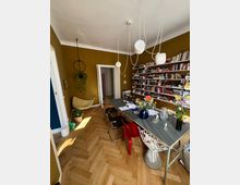 Ein stilvoll eingerichteter Raum mit gelb-oliven Wänden, einem hölzernen Fischgrätboden und einem langen Tisch mit modernen Stühlen in verschiedenen Designs. An einer Wand befinden sich zahlreiche Regale mit Büchern, während Hängelampen, eine hängende Pflanze und Vasen mit Blumen die Einrichtung ergänzen.