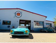 Frontansicht einer Autowerkstatt mit weißer Fassade und rotem Dachrand, großem Einfahrtstor und einem Fenster‑Rolltor an der Seite. Davor liegt ein gepflasterter Hof, auf dem Oldtimer-Fahrzeuge vor dem geöffneten Werkstatteingang stehen.