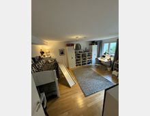 Kinderzimmer mit Holzboden, Etagenbett mit kleiner Rutsche, grauem Teppich, großen Regalen mit Spielzeug, einem Schreibtisch am Fenster und schlichten Vorhängen. Die Beleuchtung besteht aus einer Deckenlampe und einer Tischleuchte.