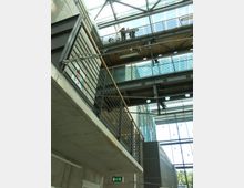 Ein modernes Gebäude-Innenraum mit sichtbaren Stahlträgern und Betonelementen. Mehrere offene Galerien mit Metallgeländern, eine gläserne Decke, die viel Tageslicht hereinlässt, und ein Notausgangsschild an der Wand sind sichtbar.