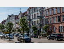 Straßenansicht mit einer Reihe von mehrstöckigen Wohnhäusern im historischen Stil, gekennzeichnet durch verzierte Fassaden, Balkone und rote Ziegelwände. Entlang der Straße stehen parkende Autos sowie kleine Bäume auf dem Gehweg.