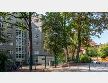 Wohngebäude aus Beton mit mehreren Etagen und großen Fenstern, umgeben von Bäumen und Grünflächen. Im Vordergrund ein Zaun, Fahrradständer sowie ein kleiner Hofbereich mit geparkten Fahrrädern und Müllcontainern.