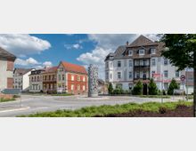 Kreisverkehr in einer städtischen Umgebung mit einem kunstvollen, metallischen Skulpturenmonument im Zentrum. Umgeben von historischen Wohngebäuden mit Satteldächern und hellen Fassaden; Schilder und Begrünung rahmen den Platz ein.