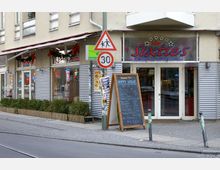 Eine Straßenszene mit einem Restaurant im amerikanischen Stil namens "The Sixties". Das Gebäude hat Glasfenster mit Neonbeschriftungen, einen Gehweg mit Pflanzkübeln sowie ein Aufsteller mit einer Speisekarte vor dem Eingang, umgeben von Verkehrsschildern, darunter ein Tempo-30-Schild.