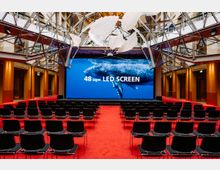 Moderner Konferenzraum mit rotem Teppich, schwarzen Stühlen in Reihen und einer großen LED-Wand mit der Aufschrift „48 sqm LED SCREEN“, auf der ein Wal abgebildet ist. Die Decke besteht aus einem Glas-Stahl-Gerüst mit weißen, wellenförmigen Dekorationen.