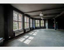 Leerer, industrieller Raum mit schwarzen Wänden und großen, symmetrischen Fenstern, durch die Tageslicht einfällt; sichtbare Heizkörper unter den Fenstern und blanker Betonboden.