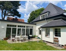 Weiße Einfamilienhaus mit dunklem Schieferdach, bodentiefen Fenstern und einer angelegten Terrasse mit weißen Gartenmöbeln. Im Vordergrund ein gepflegter Rasen, im Hintergrund vereinzelt Bäume und ein Nachbarhaus mit roten Dachziegeln.
