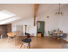 Dachgeschossraum mit schräger Decke, sichtbaren Holzbalken und drei Fenstern. Der Raum enthält Holzmöbel, darunter Tische und Stühle, eine Backsteinwand, einen Heizkörper und eine grüne Garderobe mit angebrachtem Spiegel.