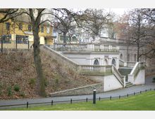 Historisches Gebäude mit weißer Fassade, verzierten Balustraden und Treppen, umgeben von kahlen Bäumen. Im Hintergrund befinden sich ein gelbes Gebäude mit Schaufenstern und weitere Stadtarchitektur.