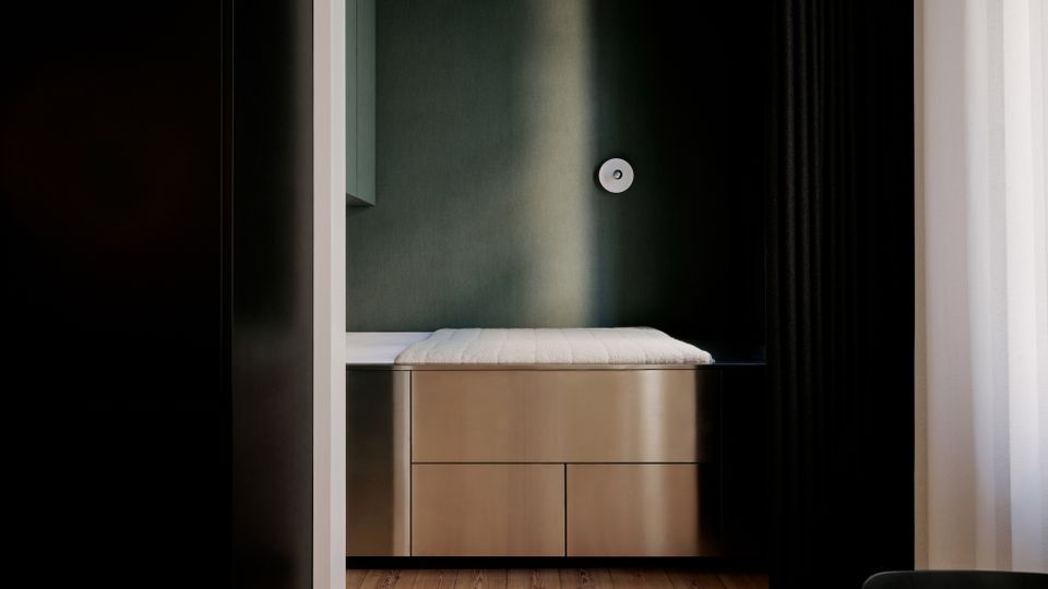 Minimalistischer Schlafbereich in einer Wandnische mit Holzfußboden, eingebautem Bett auf hellen Schubladen und einer runden Wandleuchte. Dunkle Vorhänge und ein Fenster mit transparentem Vorhang rahmen den Raum ein.