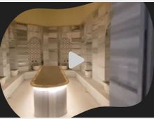 Ein traditionelles türkisches Hamam mit einer rechteckigen Marmortafel in der Mitte, umgeben von gekachelten Wänden mit dekorativen Mustern und kleinen Waschbecken. Der Raum hat eine helle Beleuchtung und eine warme, einladende Atmosphäre.