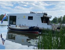 Ein modernes Hausboot mit weißem Außenbereich und der Aufschrift „The Kiss“ liegt an einem Steg auf einem ruhigen See. Im Vordergrund sind grüne Schilfpflanzen und im Hintergrund Bäume sowie weitere Boote erkennbar.