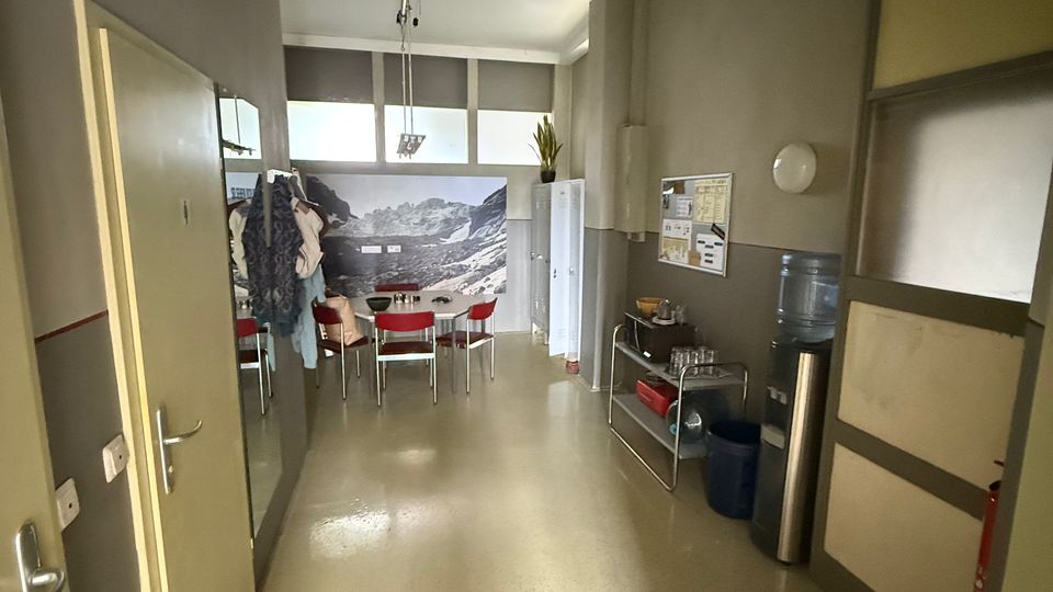 Schmaler Pausenraum in einem Büro- oder Betriebsgebäude mit grauen Wänden und hellem Boden, am Ende ein Tisch mit roten Stühlen vor einem großformatigen Bergwandbild. Rechts stehen ein Wasserspender und ein Regal mit Geschirr neben einem Aushangbrett, links sind Türen und Garderobenschränke.