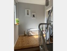 Ein kleiner, heller Flur mit Holzfußboden, grauer Wand und zwei gerahmten Bildern. Im Vordergrund ein weißes Treppengeländer, an der Wand ein Fenster mit Topfpflanze und ein cremefarbener Dekokrug.