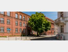 Backsteinbau mit großen, weißen Sprossenfenstern an einer Ecke einer gepflasterten Straße, flankiert von einem großen, grünen Baum. Auf der rechten Seite befindet sich ein weißes Gebäude mit Balkonelementen.