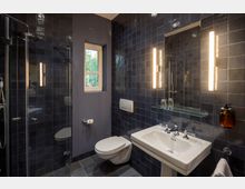 Modernes Badezimmer mit dunkelblauen Fliesen, einer Duschkabine mit Glaswänden auf der linken Seite, einem wandmontierten WC in der Mitte und einem weißen Waschbecken mit Spiegel und vertikalen Leuchten auf der rechten Seite. Ein kleines Fenster lässt Tageslicht herein, das den Raum ergänzt.