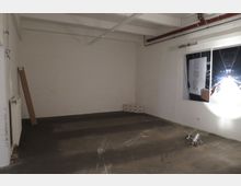 Ein leerer Raum mit weißen Wänden und einer freiliegenden Decke mit sichtbaren Rohren und Leuchten. Auf der rechten Wand hängt ein großes Poster, und auf dem Boden liegt ein einzelner Scheinwerfer.