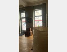 Innenansicht eines modernen Raumes mit großen Fenstern, die auf eine städtische Straße blicken. Der Raum hat Holzboden, eine hellbraune Stehlampe im Vordergrund und als Dekoration eine Anordnung aus Holzkisten sowie Kunstobjekte an den grauen Wänden.